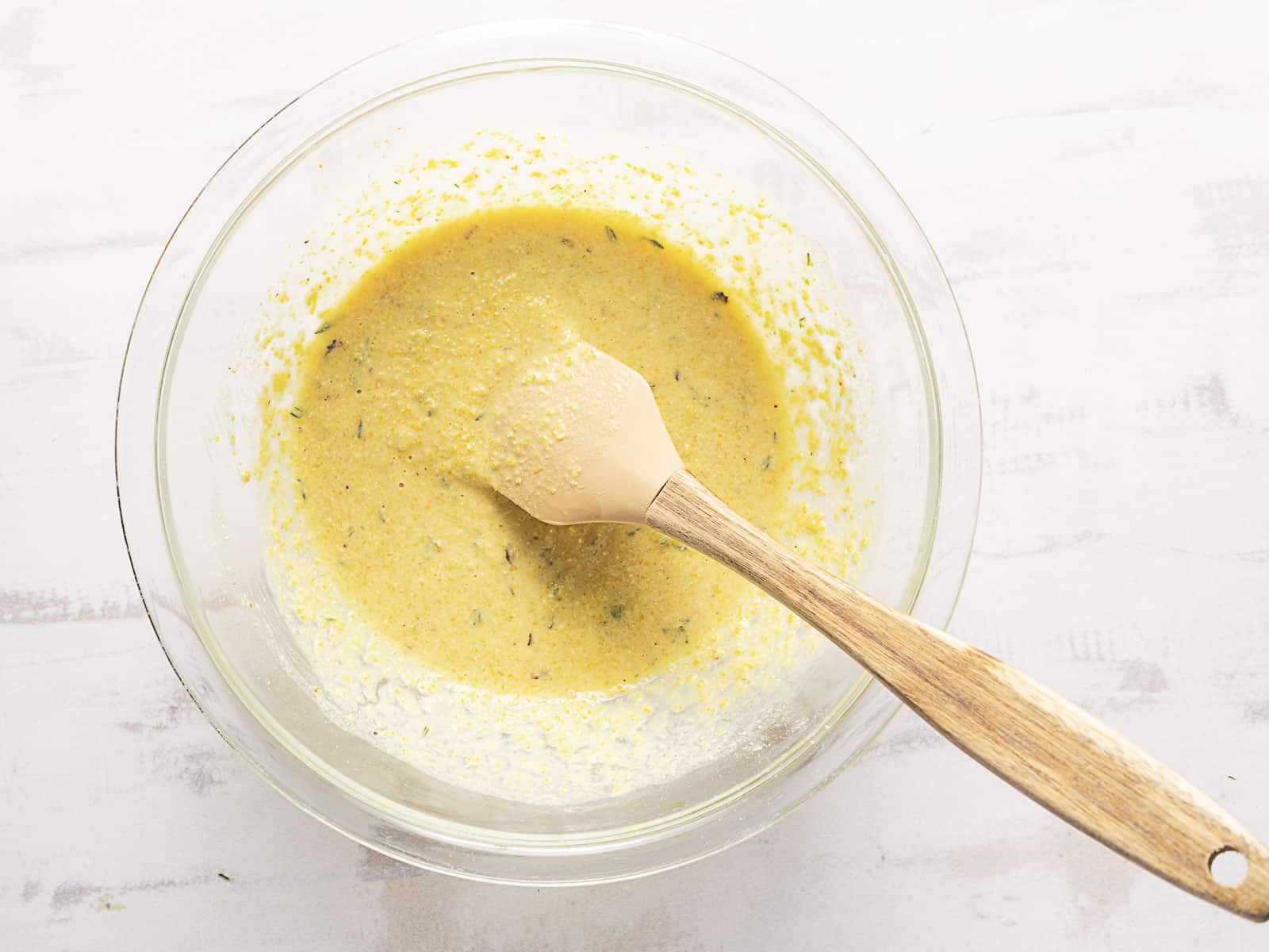 A glass bowl with yellowish batter is being mixed with a wooden spatula on a light surface.