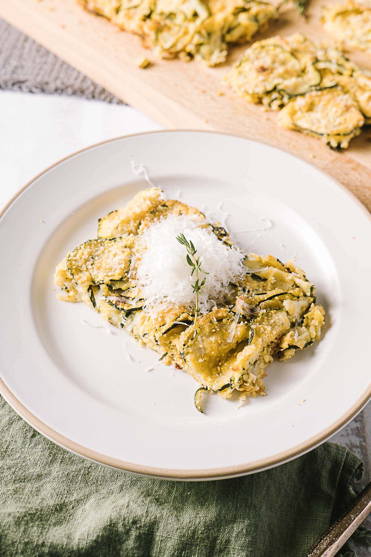 A serving of zucchini gratin with grated cheese and a thyme sprig on a white plate, placed on a green cloth. Slices of the gratin are visible on a wooden board in the background.