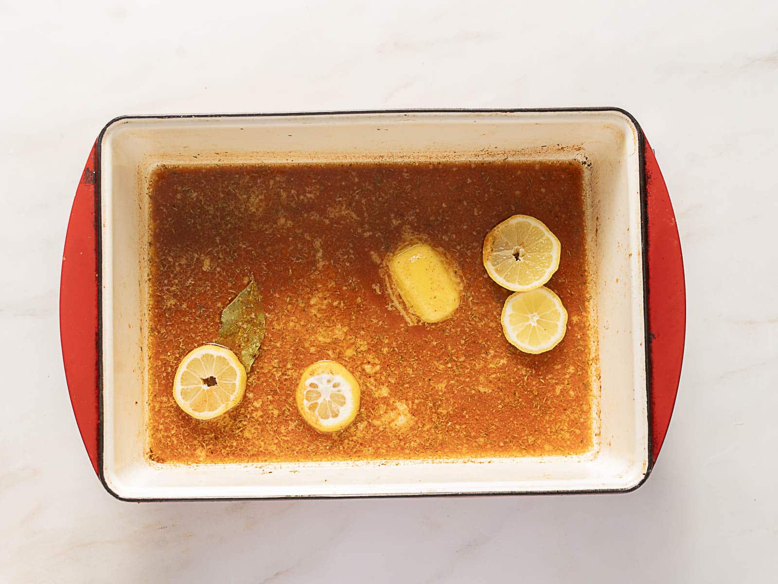 Rectangular baking dish with seasoned liquid, melted butter, lemon slices, and a bay leaf on a light surface.
