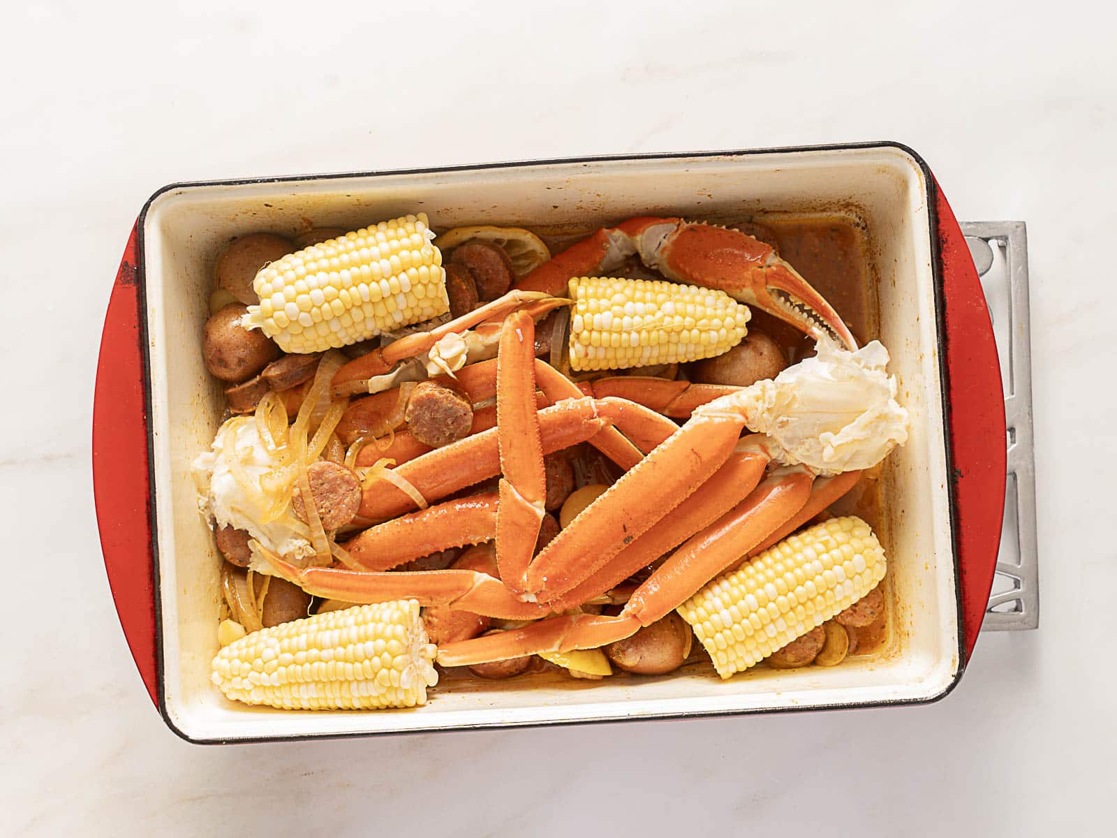 A rectangular dish filled with crab legs, corn on the cob, potatoes, sausage slices, and onions on a white surface.