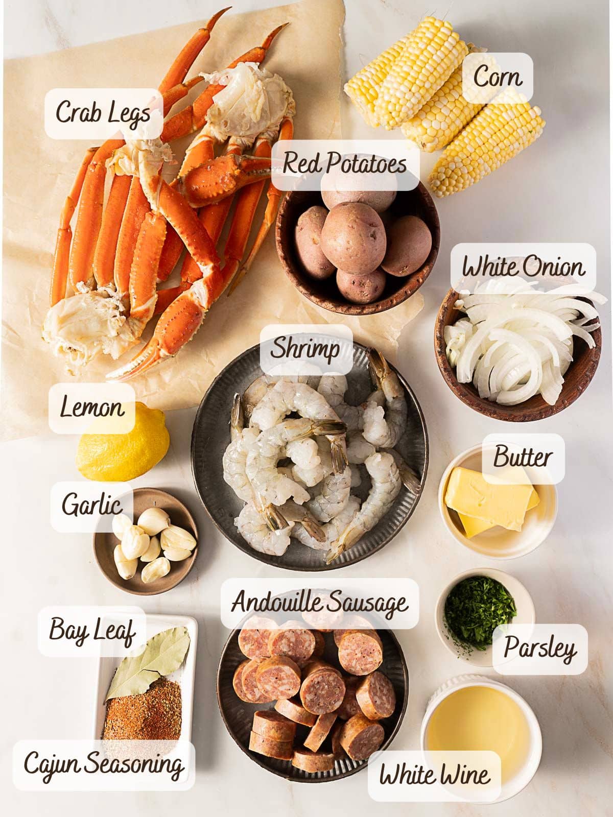 Top-down view of ingredients for a seafood bake or boil, featuring crab legs, shrimp, andouille sausage, red potatoes, corn, white onion, lemon, garlic, bay leaf, Cajun seasoning, butter, parsley, and white wine.