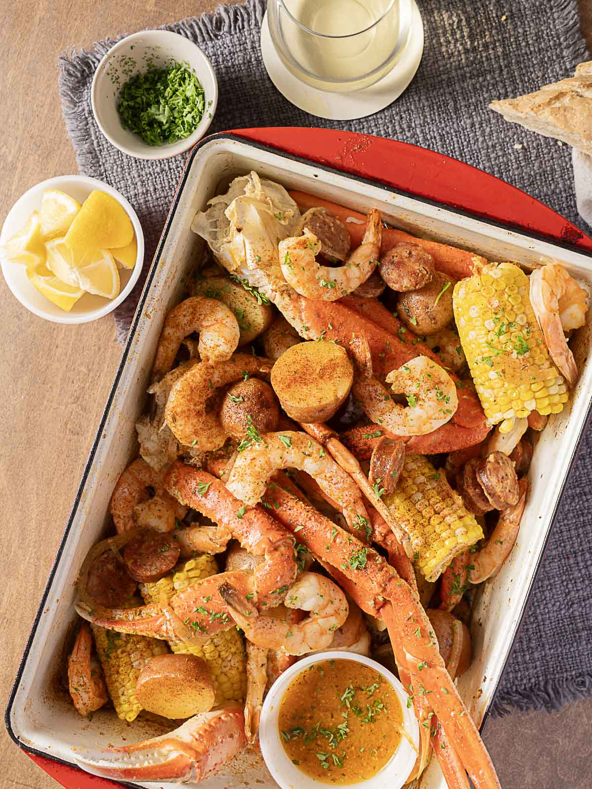 A tray of seafood boil with crab legs, shrimp, sausage, corn on the cob, and potatoes, garnished with herbs. Lemon wedges, sauce, parsley, and bread are on the side.