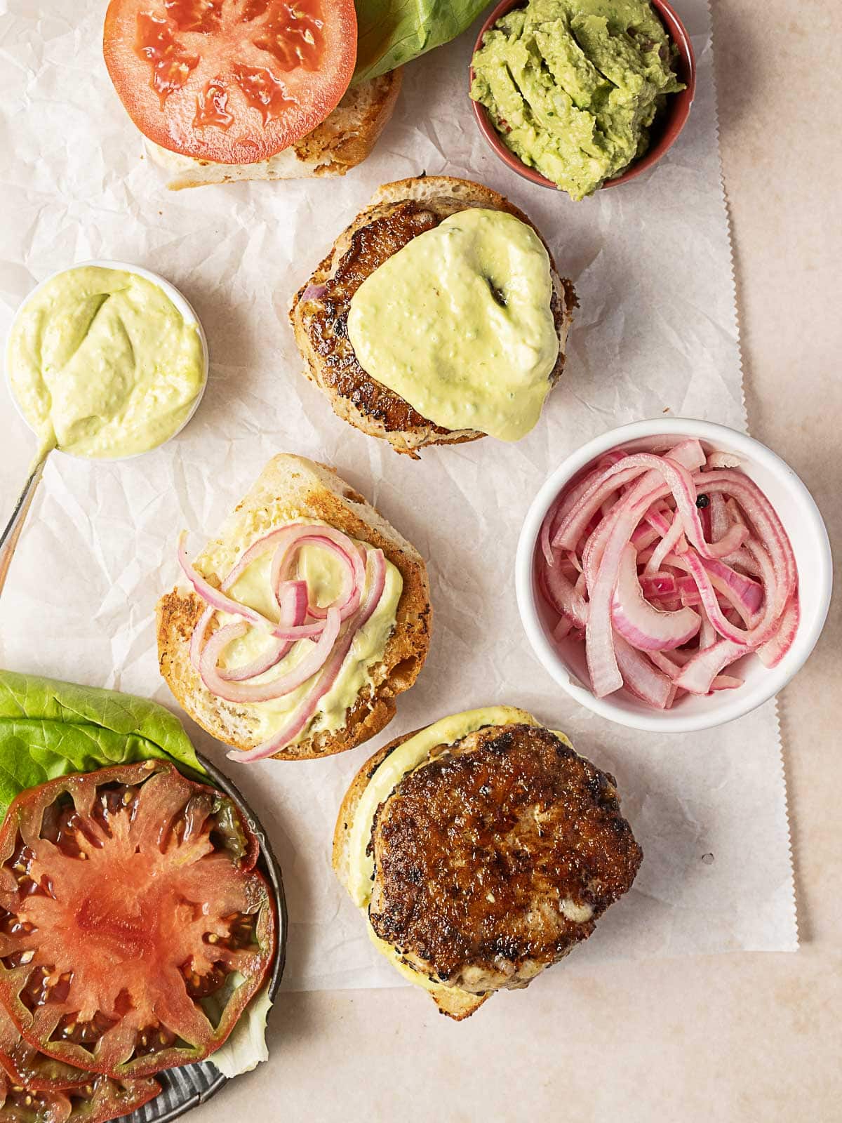 Overhead view of turkey burgers with buns, sliced tomatoes, lettuce, guacamole, pickled onions, and a spoon with a creamy sauce on parchment paper.