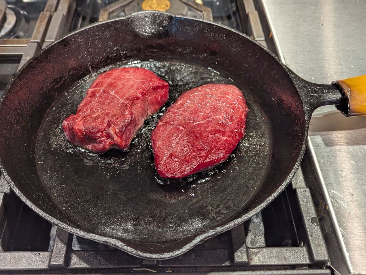 Two pieces of raw meat in a cast iron skillet on a stove.