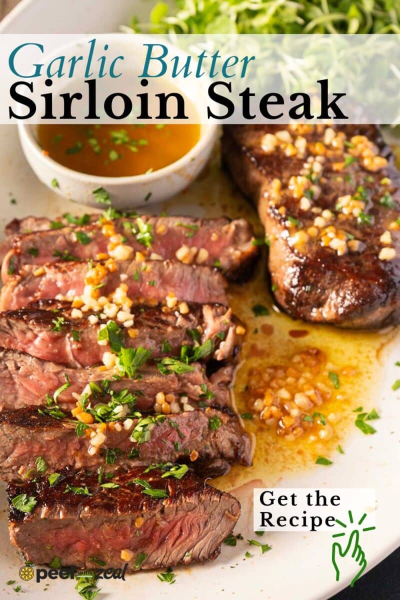 Sliced and whole sirloin steaks are generously topped with a buttery garlic spread and parsley, served on a plate with a bowl of sauce.