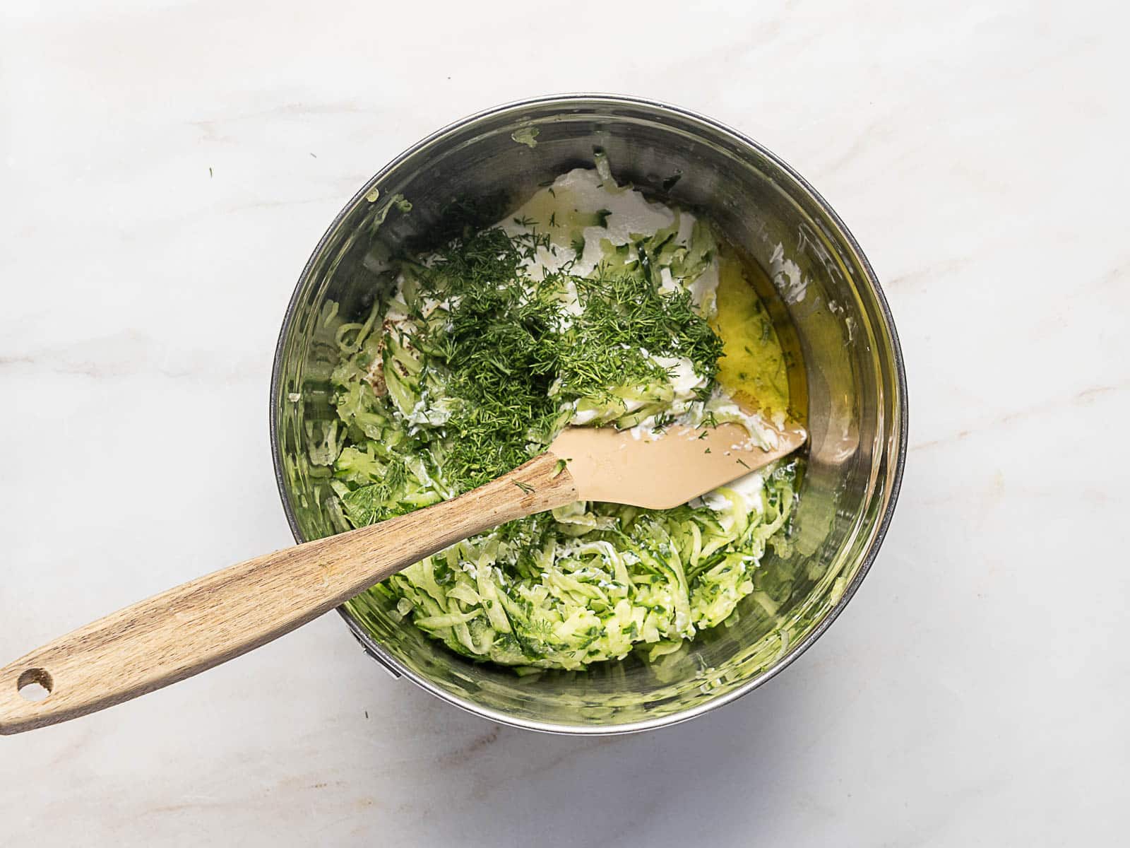 A metal mixing bowl with grated cucumber, chopped herbs, sour cream, and oil, with a wooden spatula resting inside.