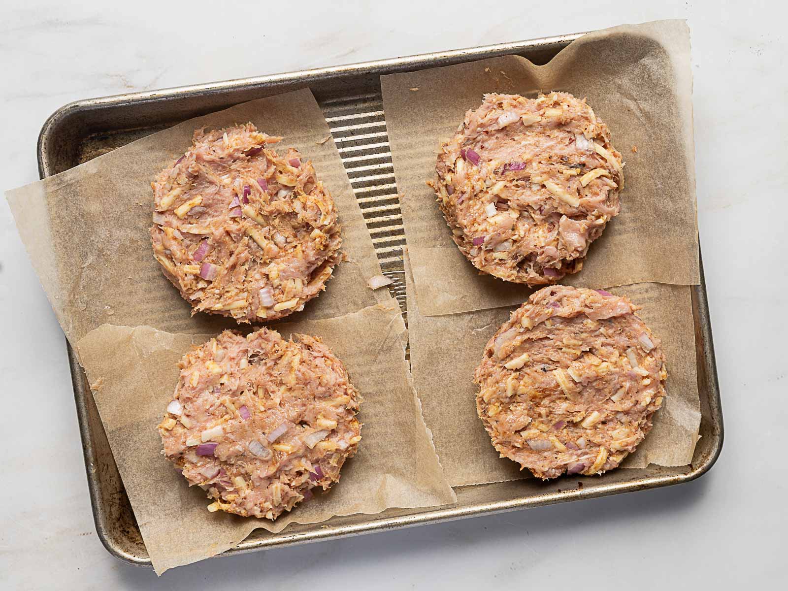 Four uncooked turkey patties with visible red onion and seasonings sit on parchment paper-lined baking tray.