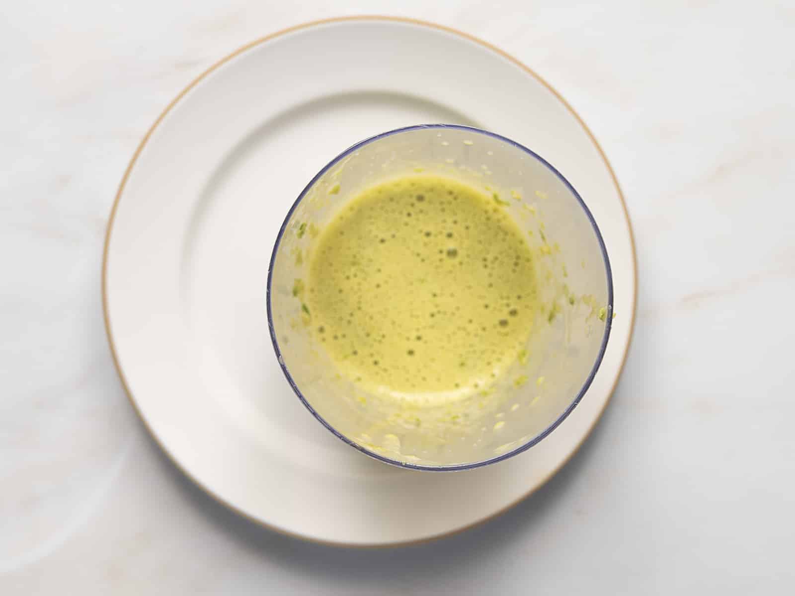 A clear cup filled with yellow-green frothy liquid sits on a white plate against a light-colored background.