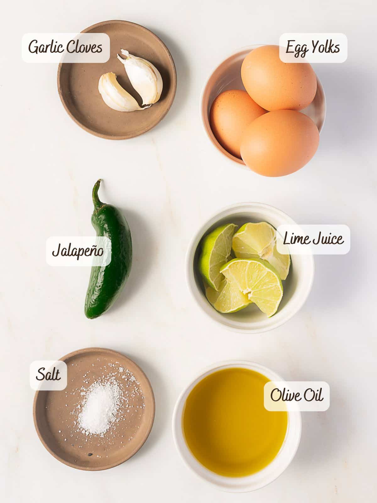 Six labeled ingredients for jalapeno mayo on a white surface: garlic cloves, egg yolks, jalapeño, lime juice, salt, and olive oil displayed in small bowls or dishes.