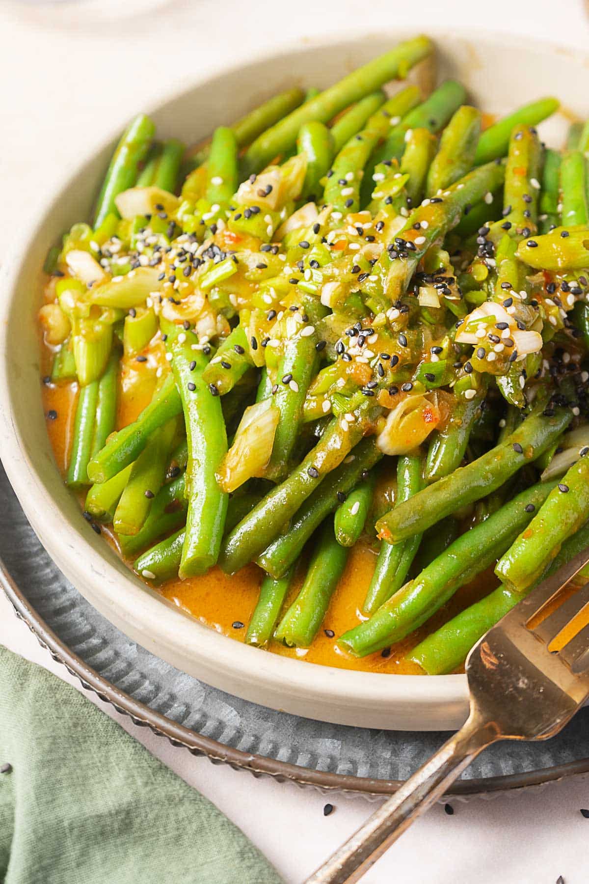 A plate of cooked green beans topped with chopped garlic, sauce, and sprinkled with black and white sesame seeds. A fork rests on the side of the plate.