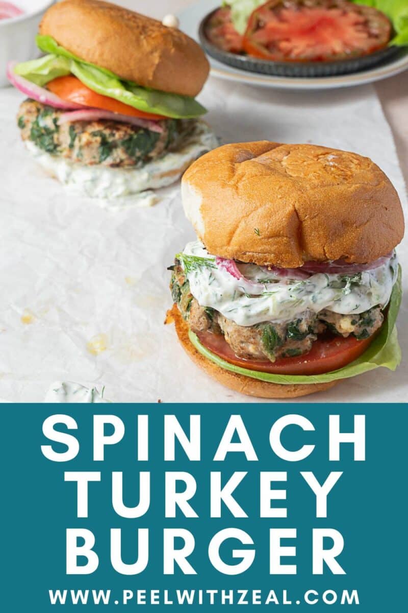 Two spinach and feta turkey burgers with lettuce, tomato, and creamy sauce on buns sit on parchment paper, with sliced tomatoes in the background. Text reads: Spinach Turkey Burger.