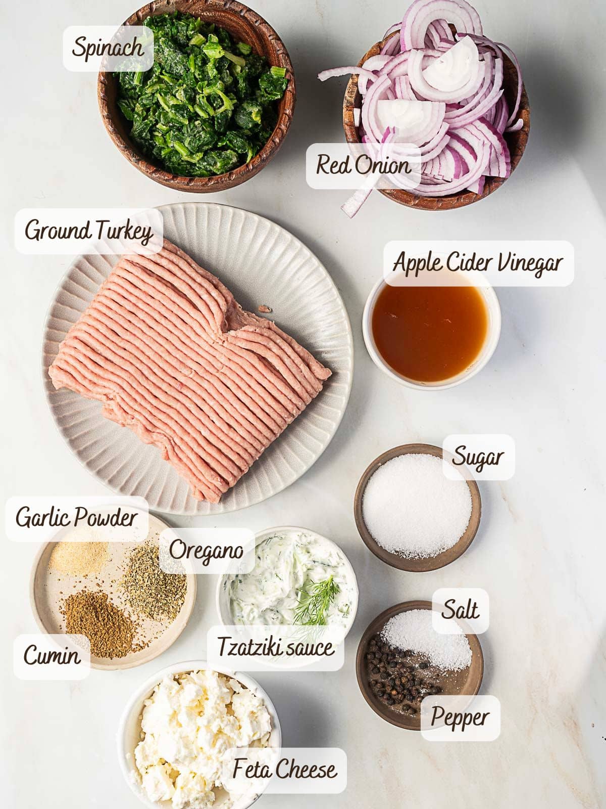 Top-down view of ingredients for spinach and feta turkey burgers, including spinach, red onion, ground turkey, apple cider vinegar, sugar, garlic powder, cumin, oregano, tzatziki sauce, feta cheese, salt, and pepper.