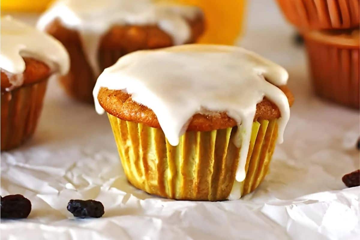 A muffin with white icing in a yellow wrapper rests on crumpled paper, evoking the charm of Easter morning. Nearby, additional muffins and a few raisins hint at delectable carrot recipes waiting to be discovered.