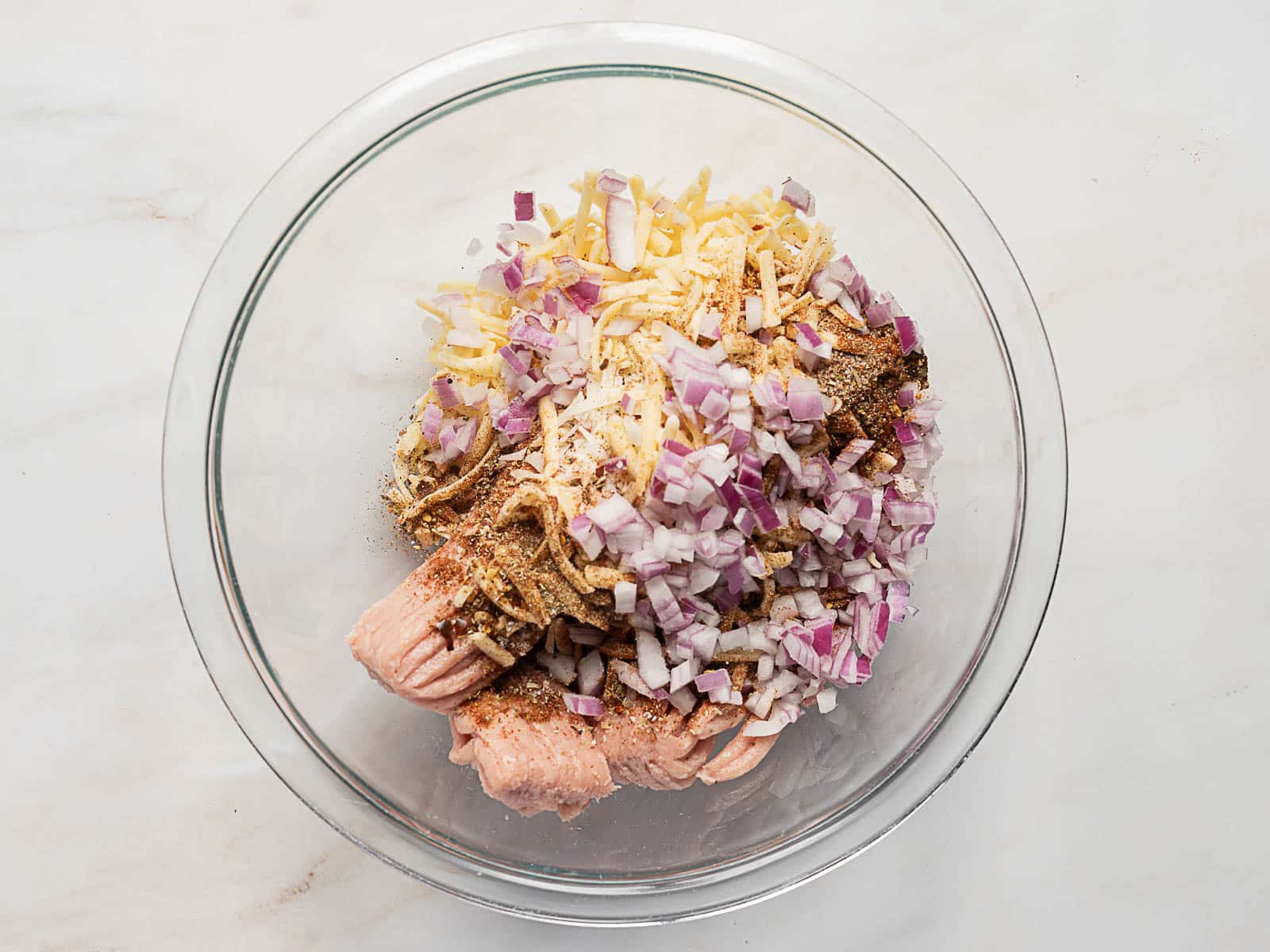 A glass bowl containing ground meat, chopped red onions, shredded cheese, and various seasonings on a light-colored surface.