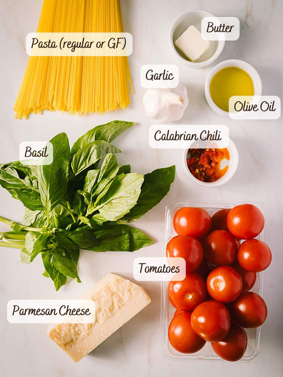 Various fresh ingredients for Tomato Basil Pasta are arranged on a surface, including pasta, butter, olive oil, garlic, Calabrian chili, basil, tomatoes, and Parmesan cheese—each ingredient clearly labeled.