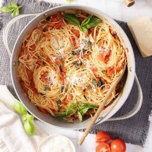 A pot of cooked spaghetti with tomato sauce, basil leaves, and grated cheese, with a wooden spoon resting inside. Fresh tomatoes, basil, and cheese are nearby.