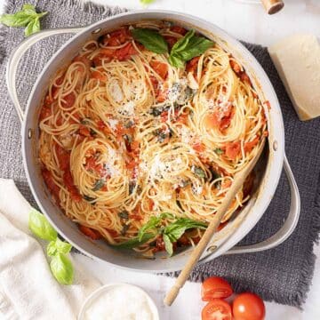 A pot of cooked spaghetti with tomato sauce, basil leaves, and grated cheese, with a wooden spoon resting inside. Fresh tomatoes, basil, and cheese are nearby.