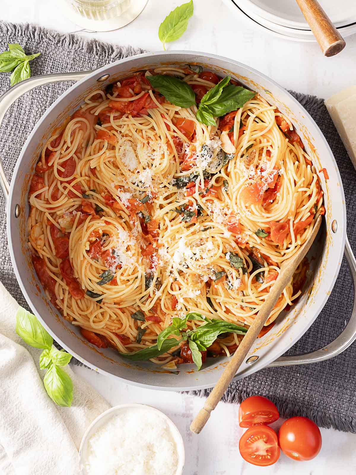 A pot of cooked spaghetti with tomato sauce, fresh basil, and grated cheese, with a wooden spoon resting inside. Cherry tomatoes and a bowl of grated cheese are nearby.