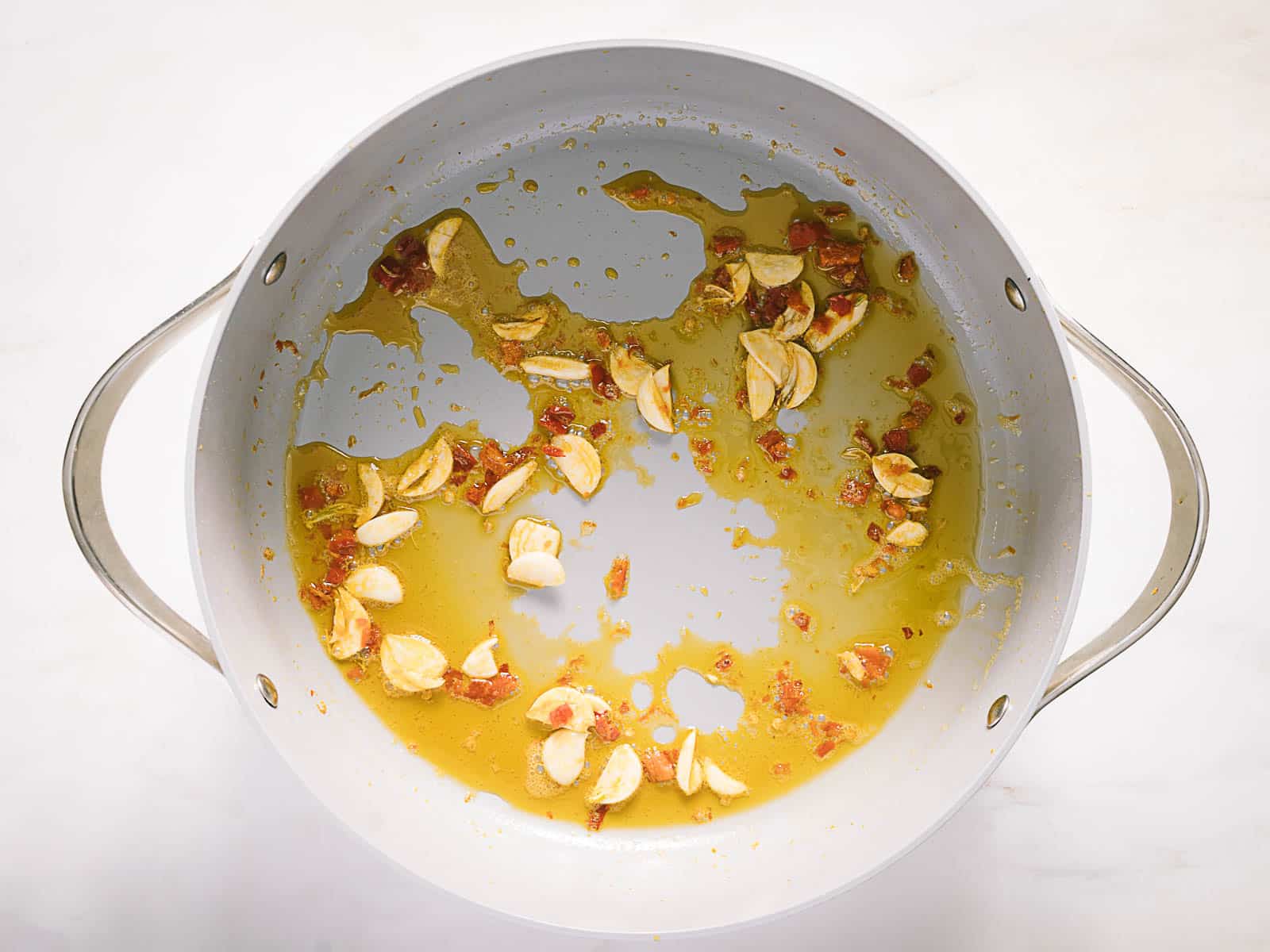 A white pan with thinly sliced garlic and small bits of red chili frying in oil on a white surface.