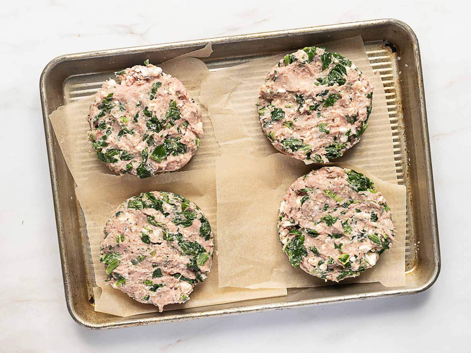 Four raw turkey burger patties with spinach and feta are arranged on parchment paper squares on a metal baking sheet.