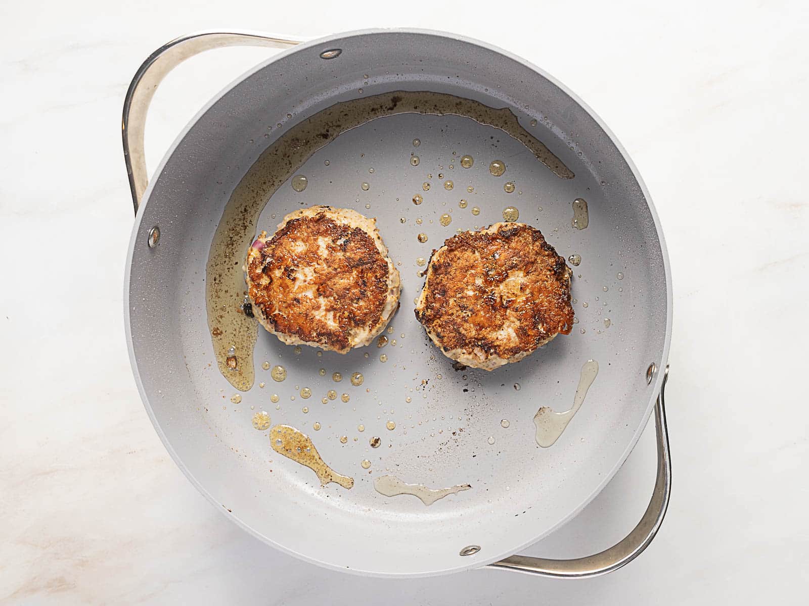 Two browned patties cooking in a light-colored frying pan with some oil, viewed from above.