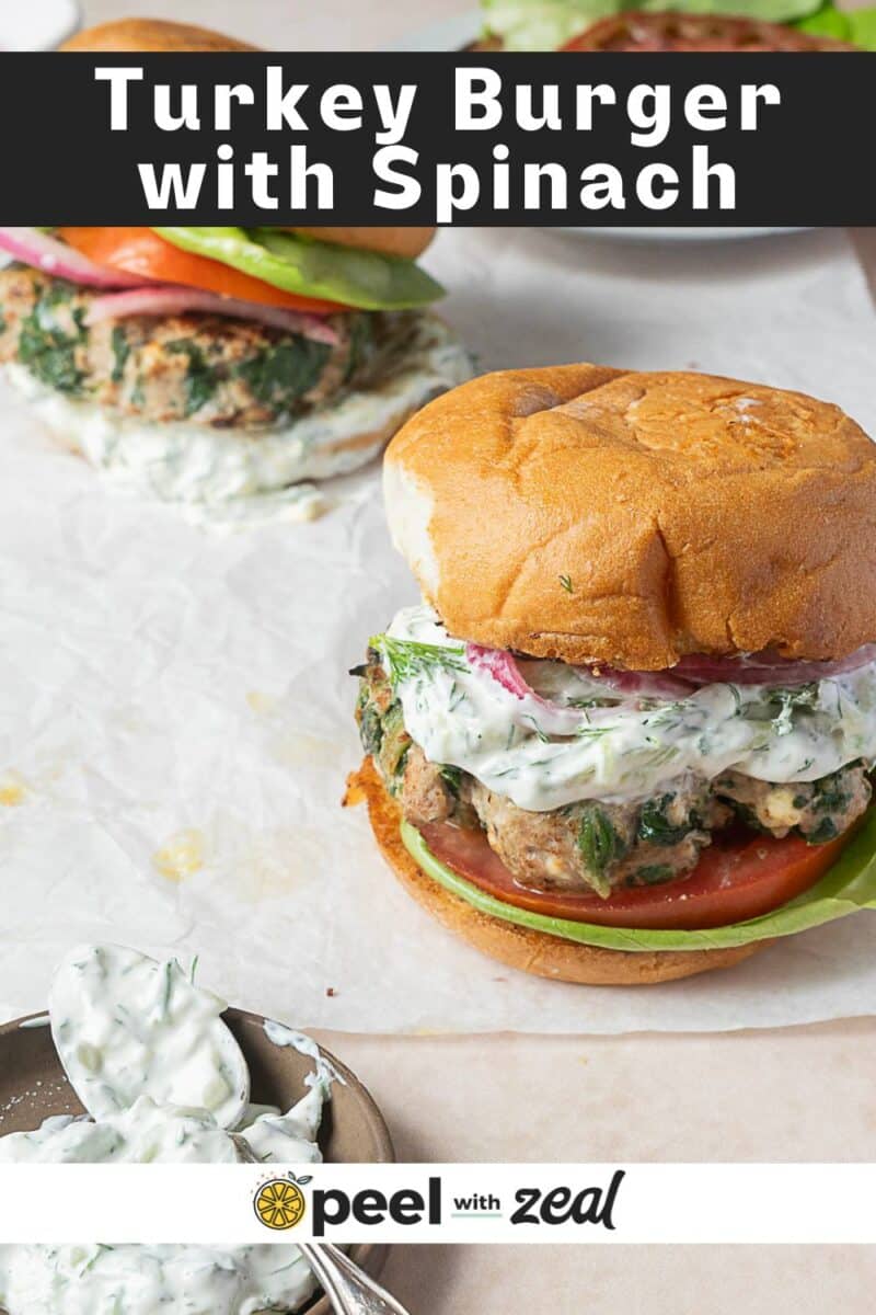 A spinach and feta turkey burger with tomato, red onion, lettuce, and creamy sauce on a bun sits on parchment paper, with a second burger and a bowl of sauce nearby.