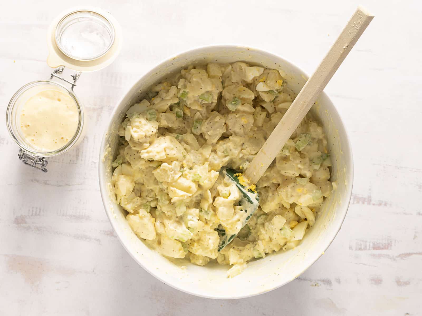 Bowl of potato salad with a wooden spoon on a light surface, next to a jar of dressing.
