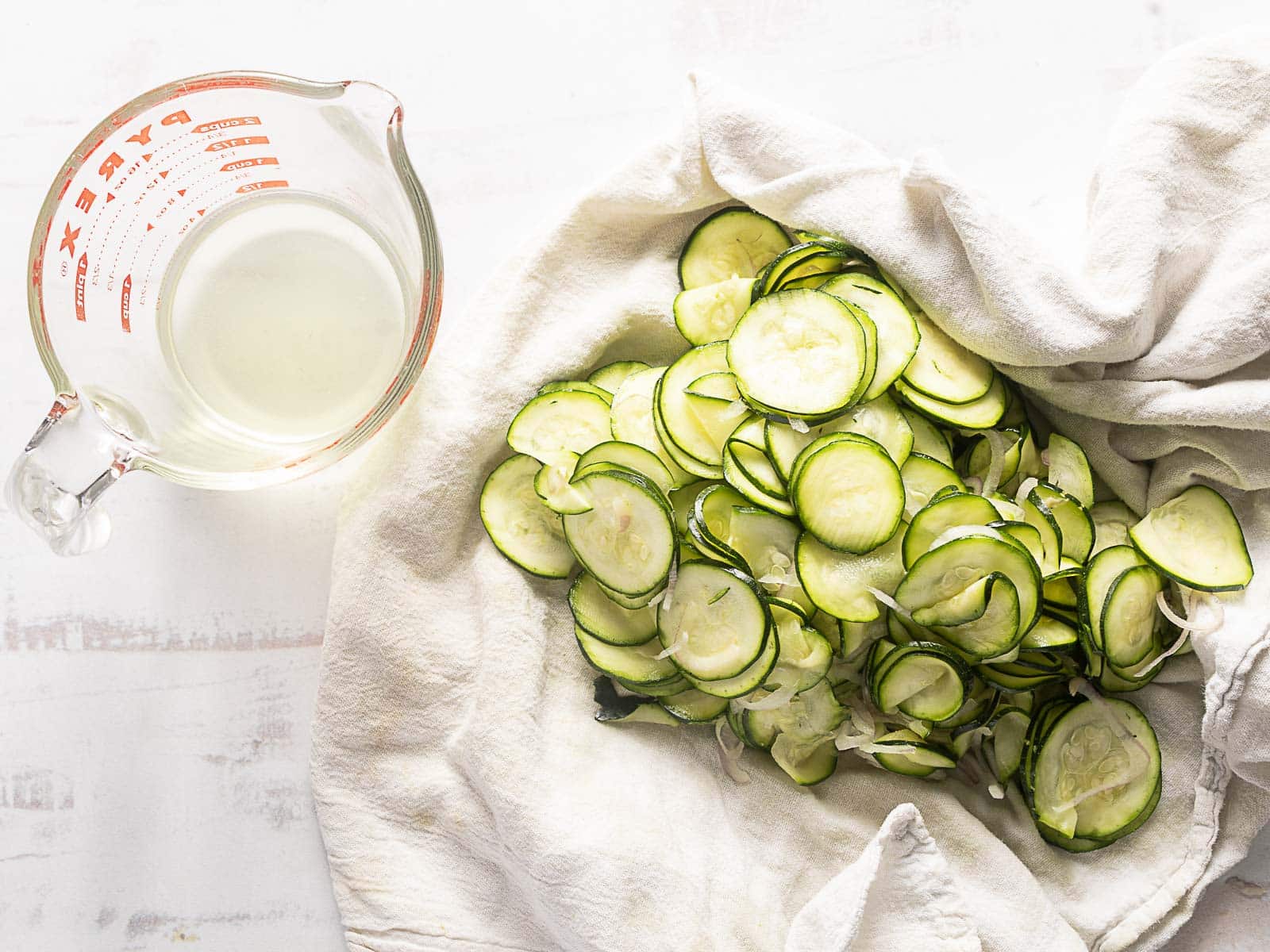 Sliced cucumbers on a white cloth next to a Pyrex measuring cup on a white surface.