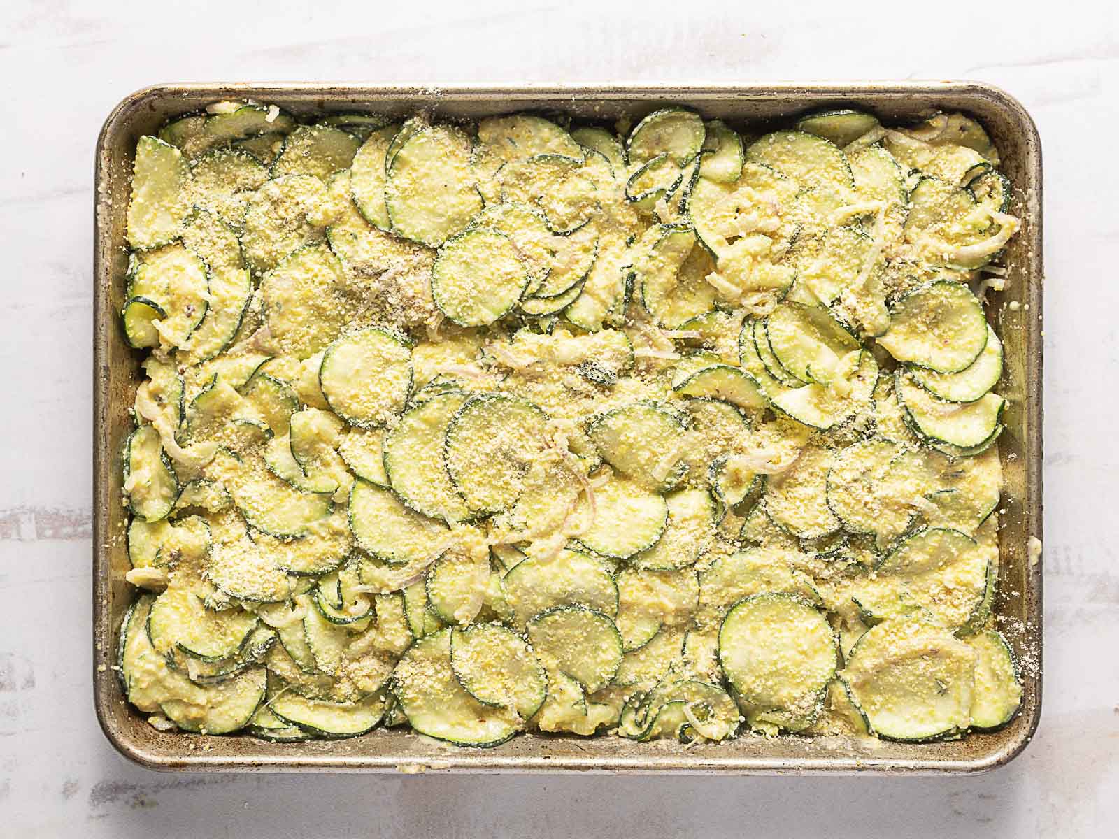 A rectangular baking dish filled with sliced zucchini, topped with grated cheese and bread crumbs, ready for baking.