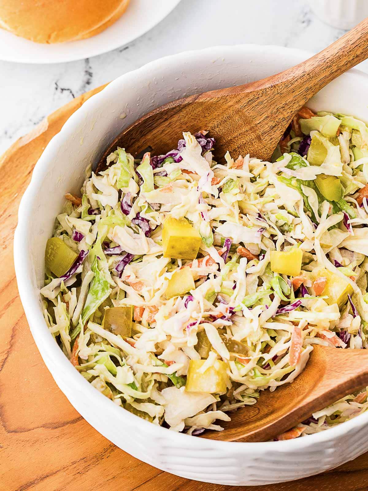 A bowl of coleslaw with shredded cabbage, carrots, pickles, and a creamy dressing, with two wooden serving utensils.