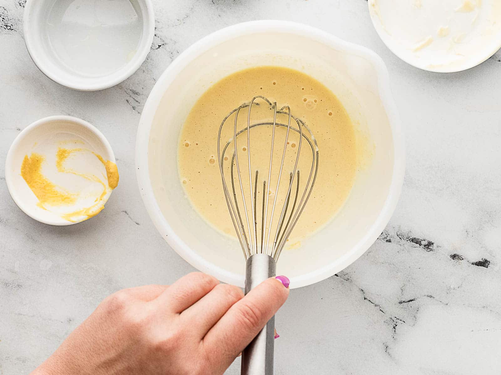 A hand holding a whisk mixes a yellow batter in a white bowl on a marble countertop, with small bowls nearby.