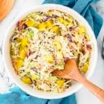 Bowl of coleslaw with shredded cabbage, carrots, purple cabbage, and yellow diced pickles, mixed with creamy dressing. Wooden spoon rests in the bowl.