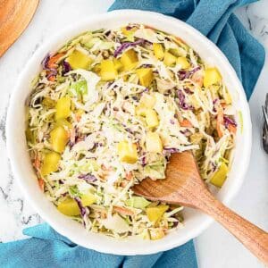 Bowl of coleslaw with shredded cabbage, carrots, purple cabbage, and yellow diced pickles, mixed with creamy dressing. Wooden spoon rests in the bowl.