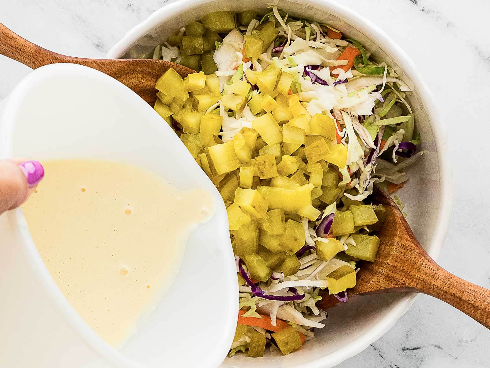 A bowl of coleslaw with chopped pickles is being prepared; creamy dressing is being poured in while two wooden spoons rest in the bowl.