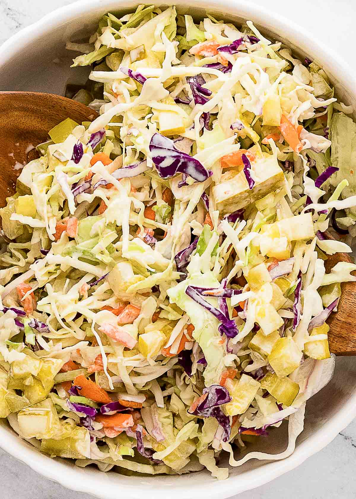 A bowl of coleslaw with shredded green and purple cabbage, carrots, pickles, and apple pieces, mixed with dressing and served with wooden salad utensils.