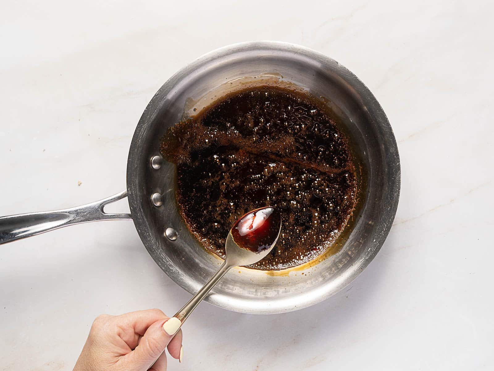 A hand holds a spoon over a stainless steel pan with a dark brown sauce simmering inside, set on a white countertop.