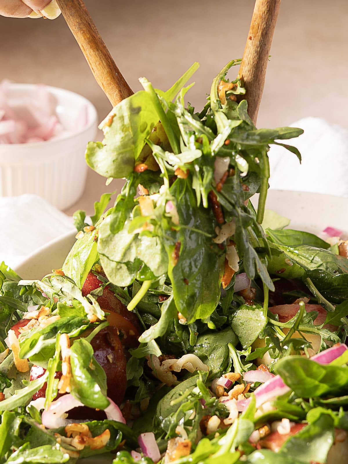 A close-up of a mixed green salad with red onions and nuts being tossed with wooden salad servers.