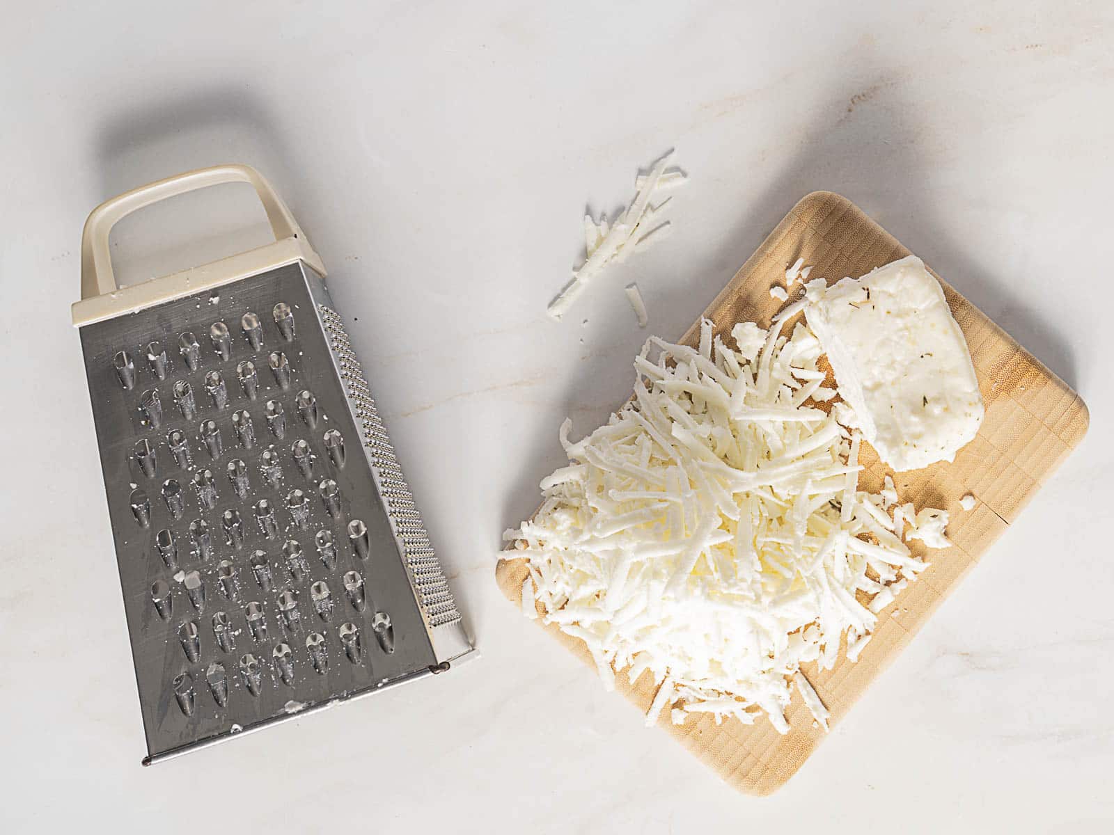 A metal box grater next to a wooden cutting board with a block of white cheese and a pile of freshly grated cheese.