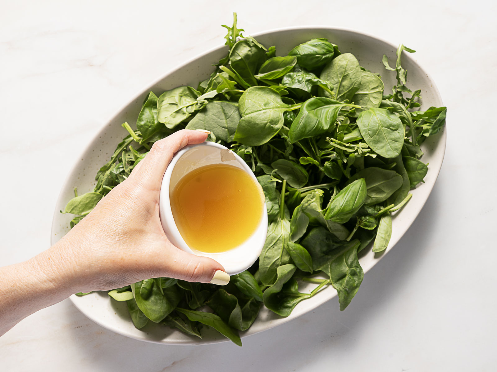 A hand holds a small bowl of vinaigrette over a large white plate filled with fresh spinach and arugula leaves.