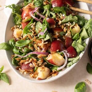 A salad with greens, sliced nectarines, red onions, toasted rice, and fresh basil leaves arranged in an oval dish.