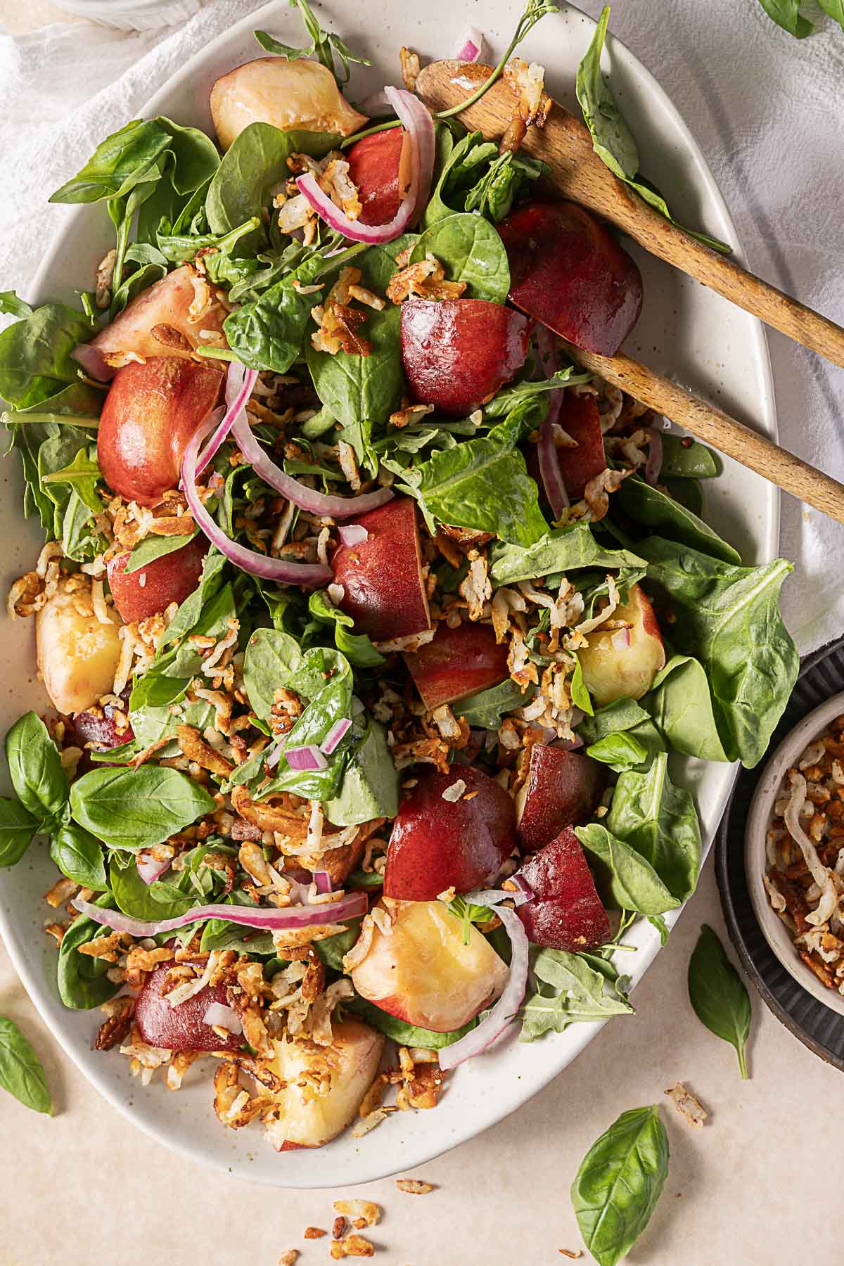 A salad with spinach, sliced red onions, rice, fresh basil, crispy fried onions, and pieces of red and yellow stone fruit on a white oval plate with wooden salad tongs.