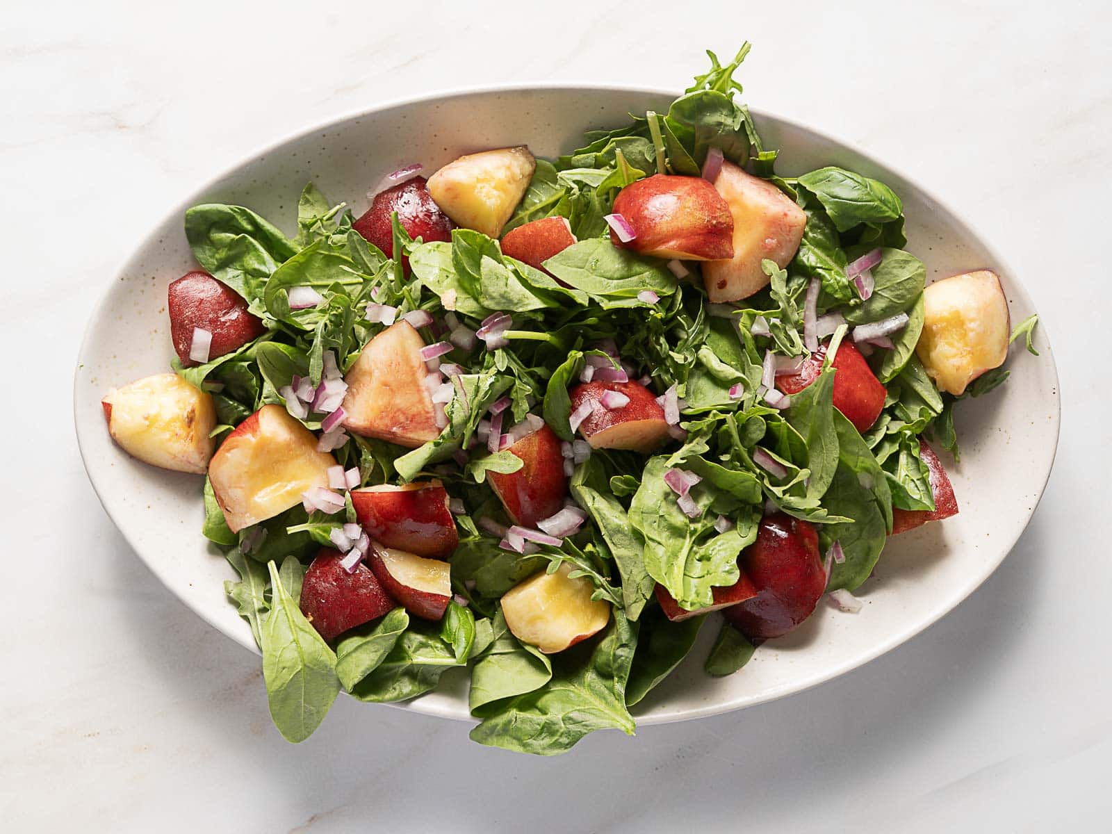 A white oval plate with a salad of spinach, arugula, sliced peaches, plums, and chopped red onions on a white surface.