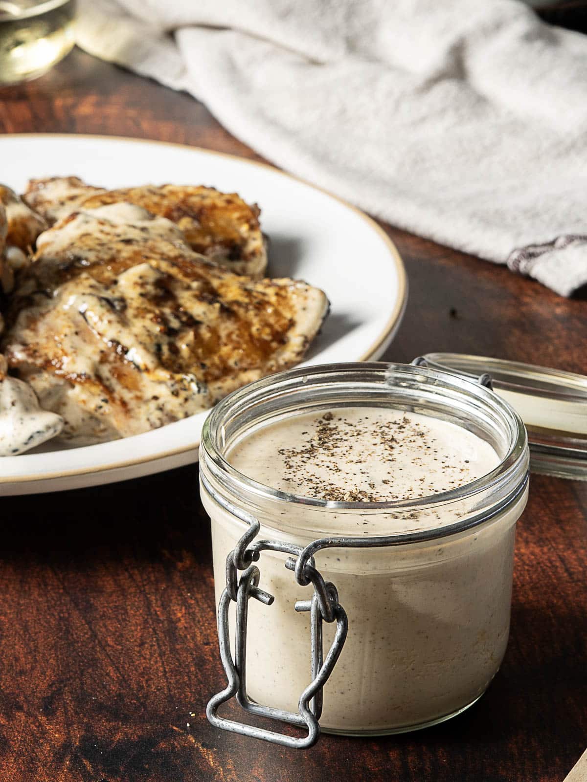 A glass jar filled with creamy white sauce topped with black pepper sits in front of a plate of grilled chicken on a wooden table.