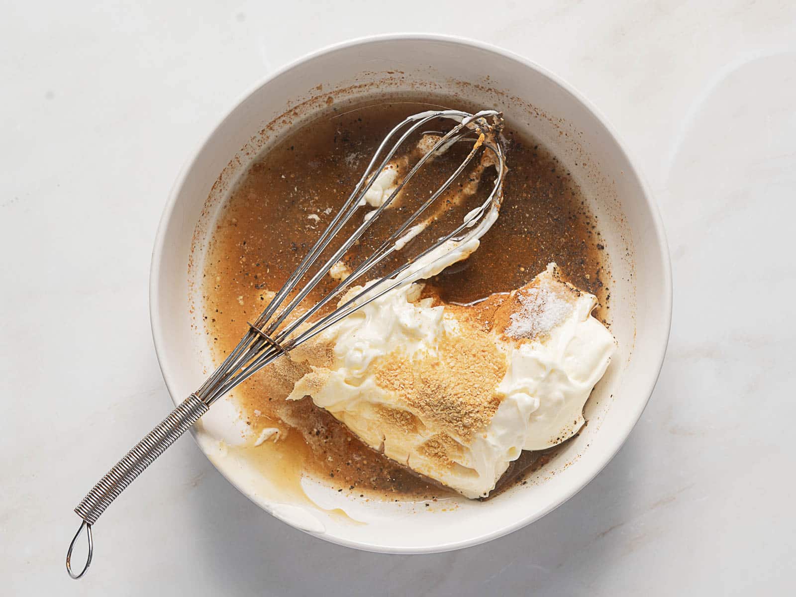 A bowl with a whisk contains cream cheese, brown sugar, and spices being mixed together on a light countertop.