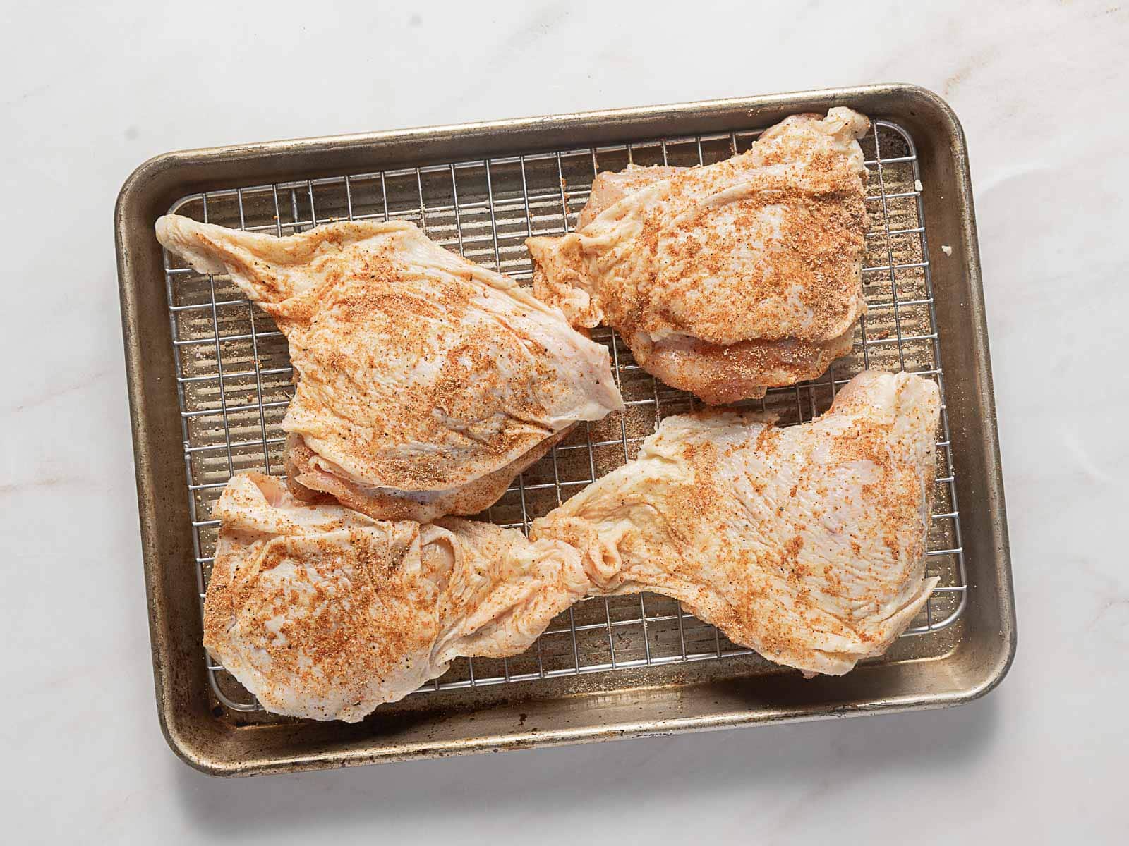 Four raw, seasoned chicken quarters on a wire rack placed over a baking tray, ready for cooking.