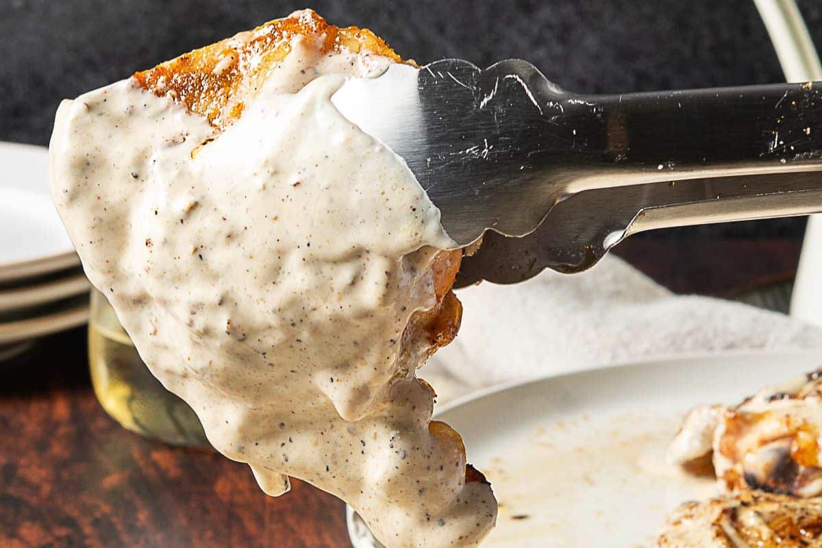 A metal tong holds a piece of fried chicken covered in white pepper gravy above a plate.