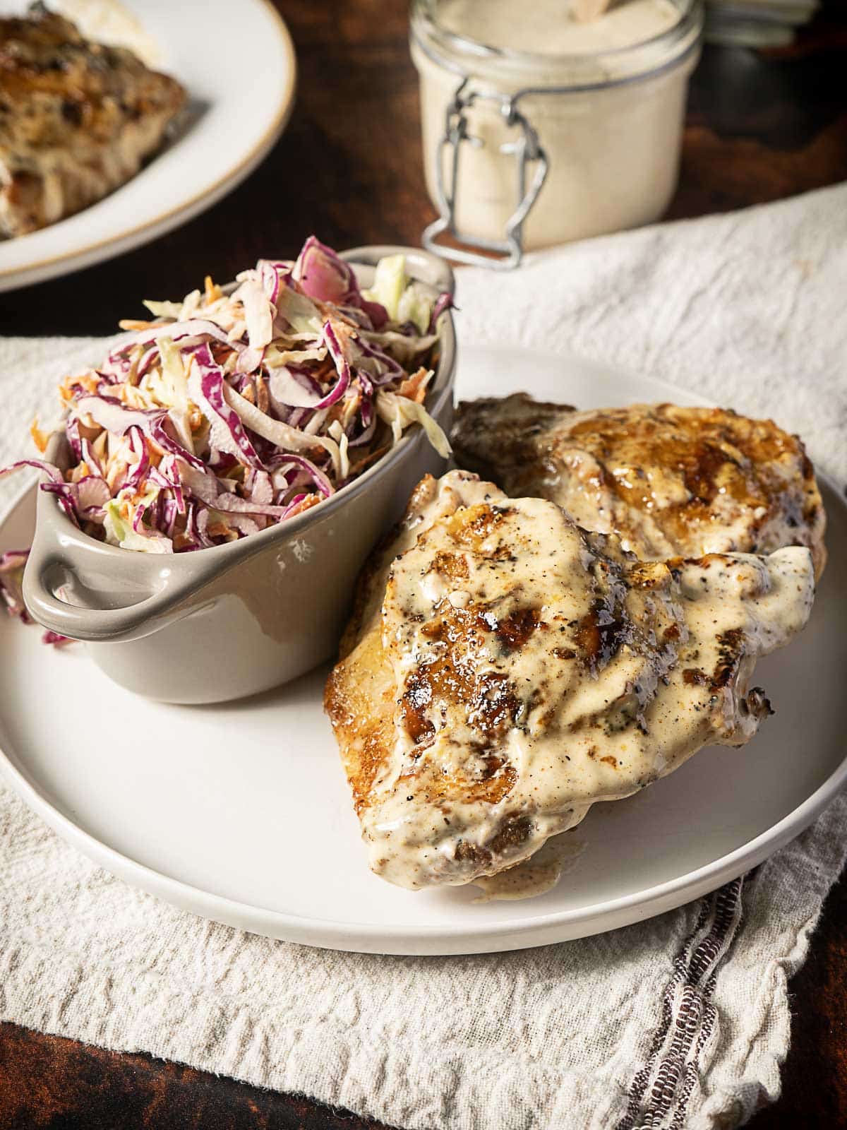 A plate with grilled chicken topped with a creamy sauce, served beside a small dish of coleslaw on a white cloth.