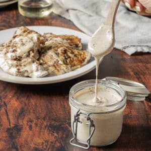 A jar of creamy white sauce is being spooned, with a plate of grilled chicken covered in the same sauce in the background.