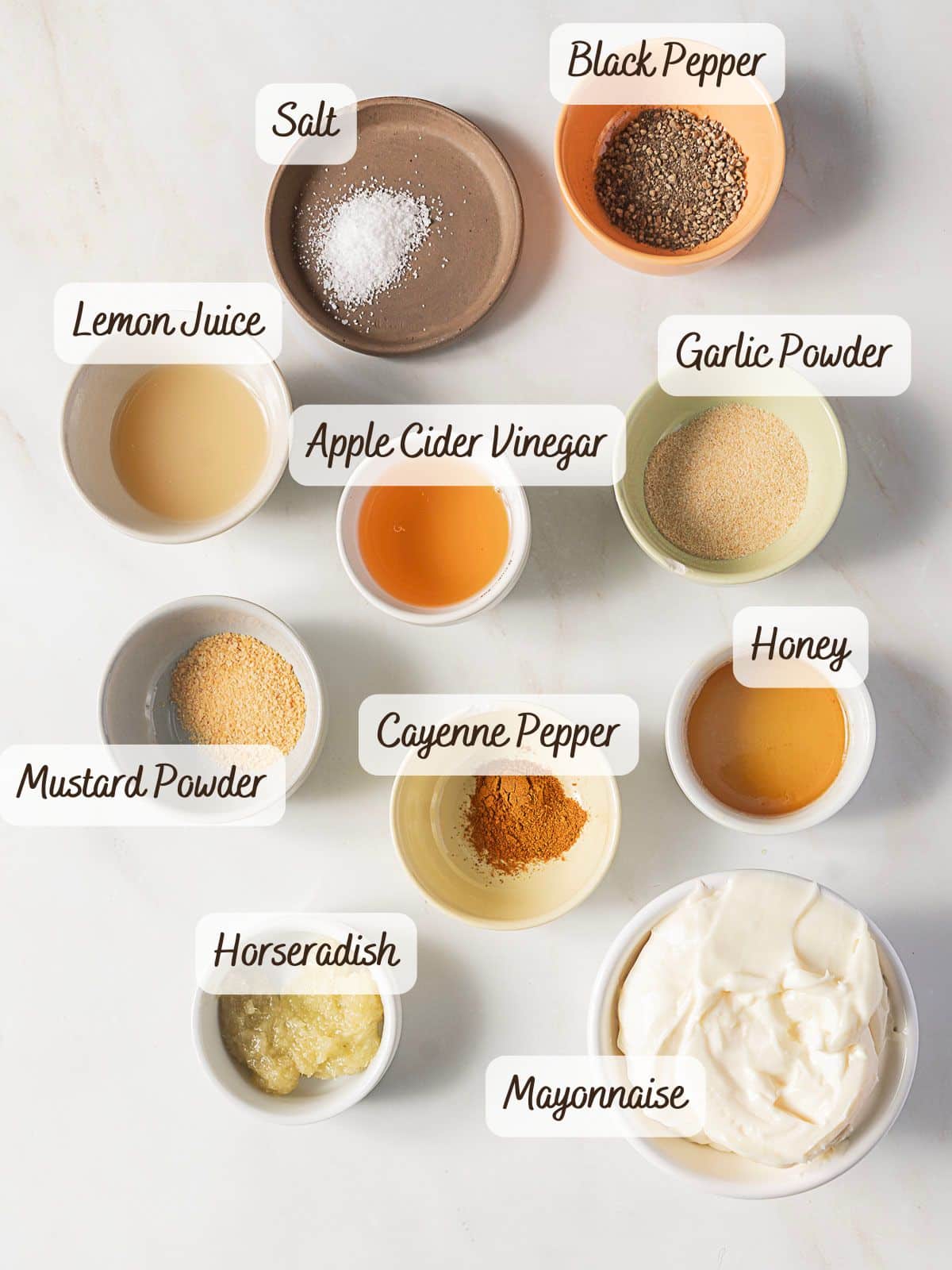 Various small bowls containing ingredients like salt, black pepper, lemon juice, apple cider vinegar, garlic powder, honey, mustard powder, cayenne pepper, horseradish, and mayonnaise—perfect for making Alabama white sauce—are arranged on a white surface.