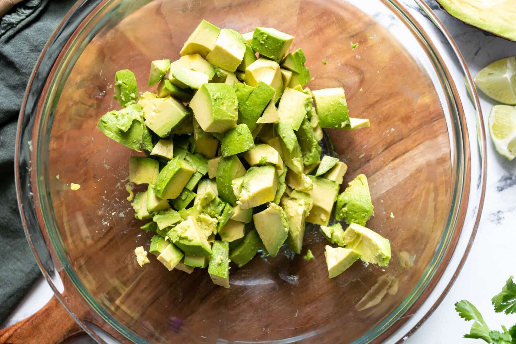 Chopped avocado pieces in a glass bowl on a wooden surface, with lime and cilantro visible nearby.